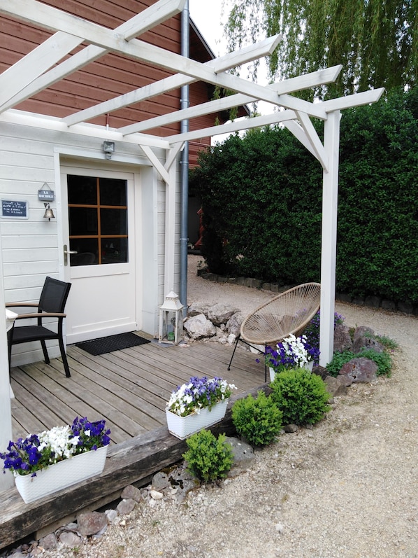 Una terraza de madera con una pérgola, una silla y macetas de flores.