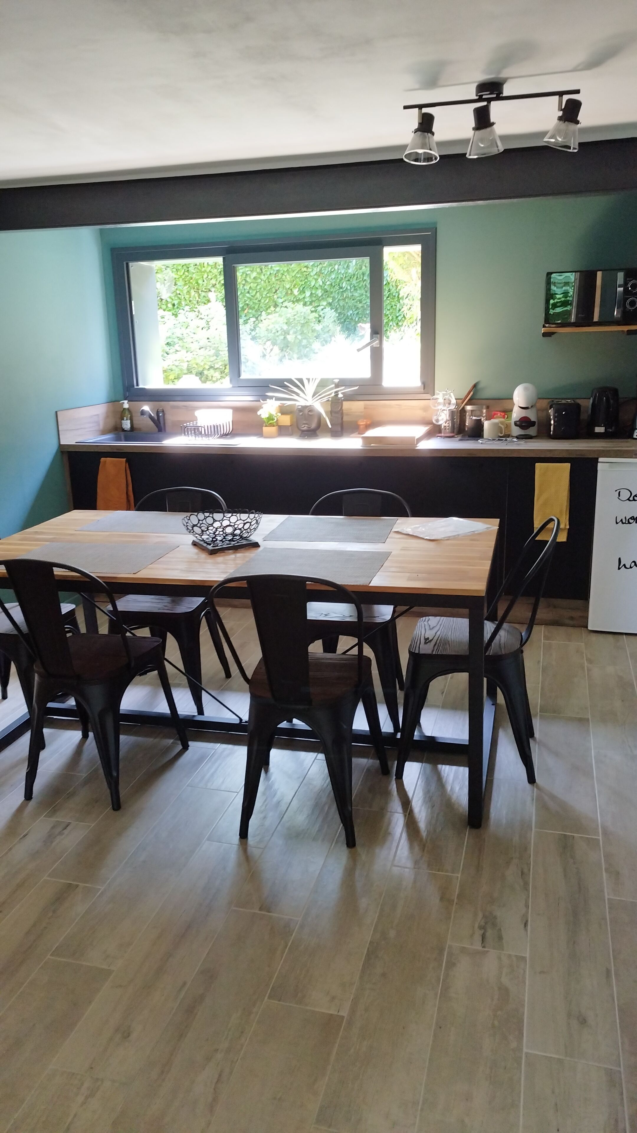 A modern kitchen with a dining area, featuring a wooden table and chairs, a large window with a view of greenery, and a built-in oven.