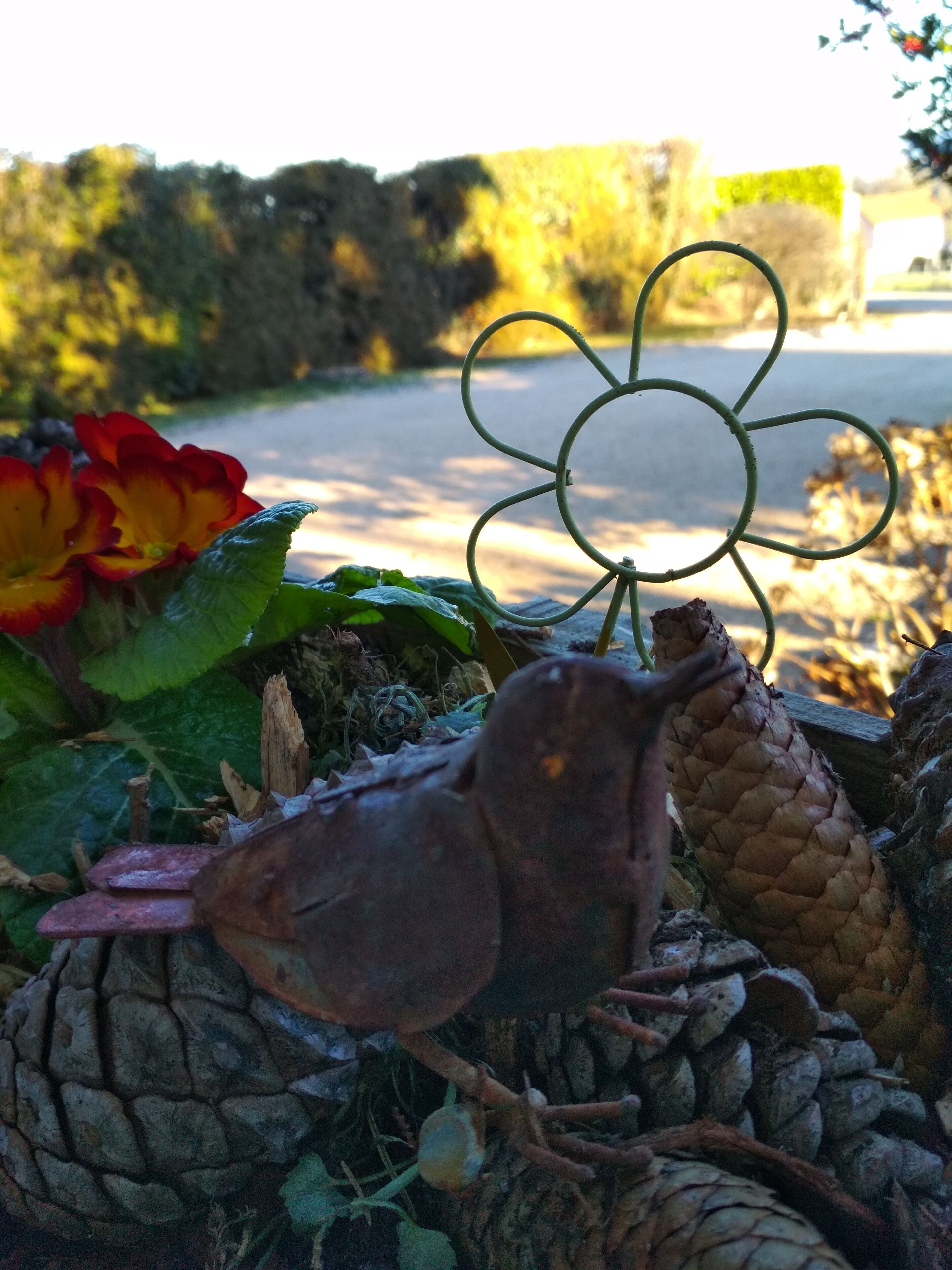 Primer plano de un objeto metálico con forma de flor, posiblemente una decoración de jardín, rodeado de elementos naturales como hojas y una piña.