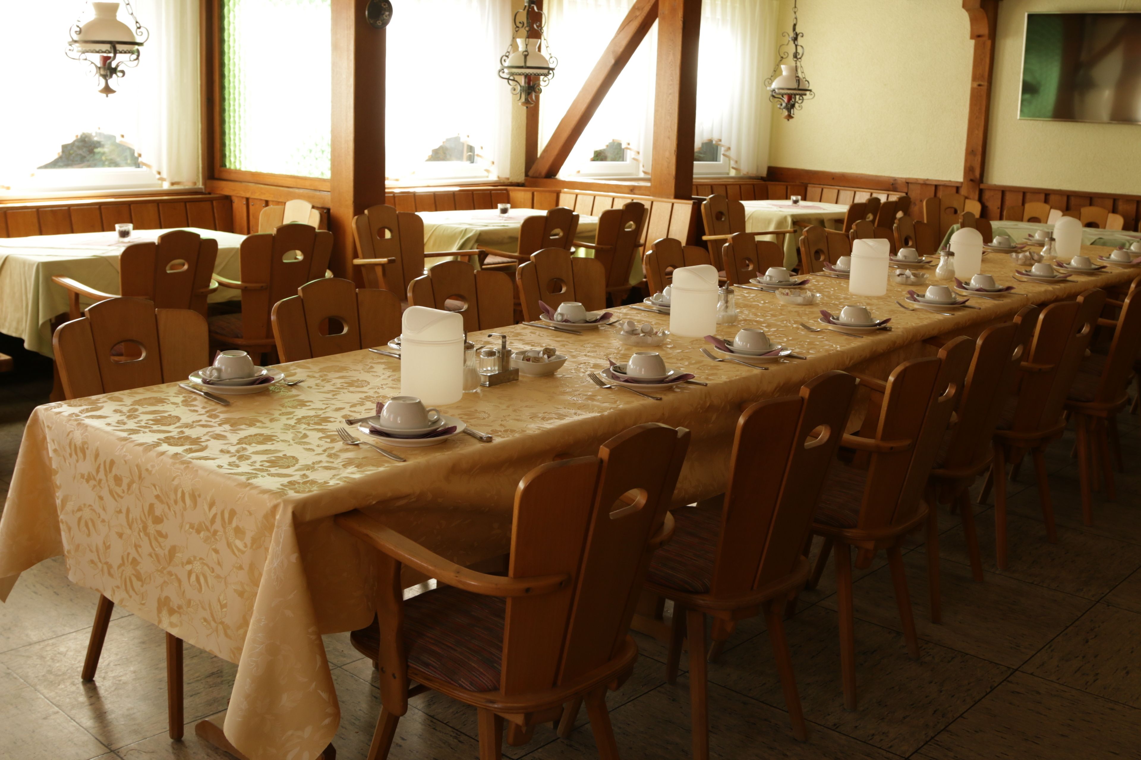 A long dining table set with chairs, tables, and hanging light fixtures in a room with windows and wooden beams.