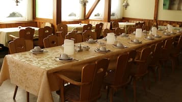A long dining table set with chairs, tables, and hanging light fixtures in a room with windows and wooden beams.
