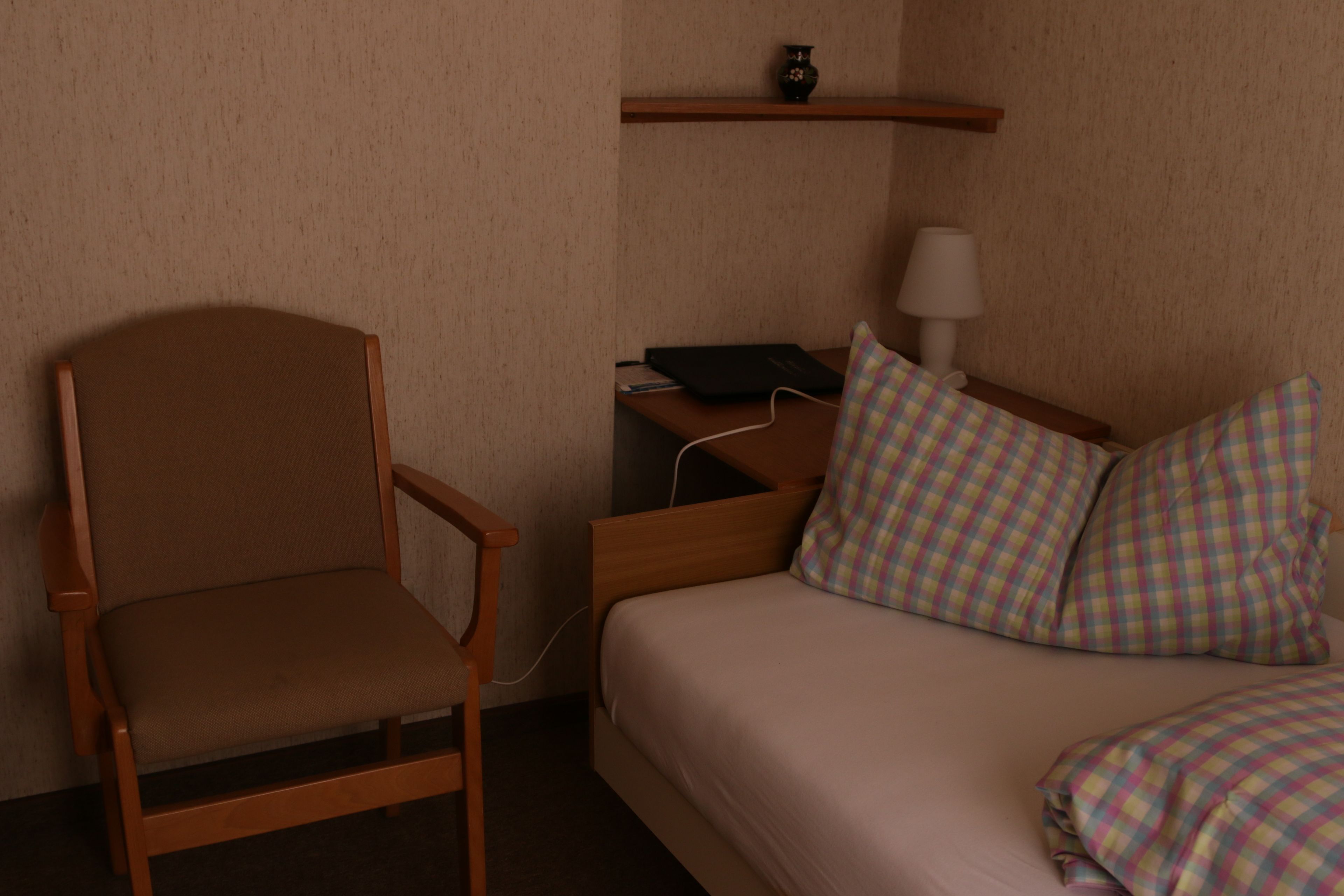 Ein Hotelzimmer mit einem Holzstuhl, einem Bett mit Karo-Bettwäsche, einem Nachttisch mit Lampe und einem Regal über dem Bett.