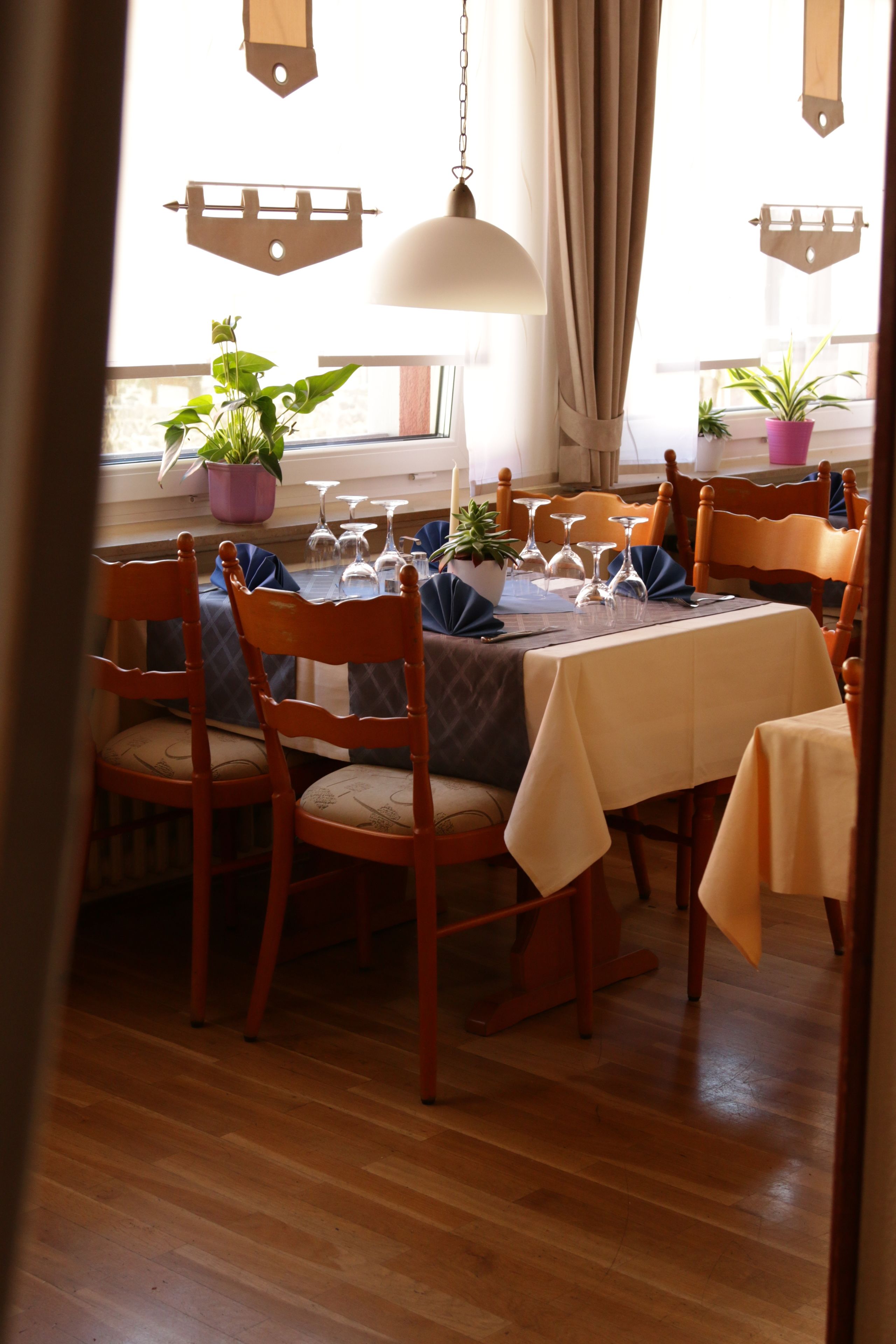 Ein Essbereich mit Holzkstühlen, einem Tisch mit weißer Tischdecke, Topfpflanzen und hängenden Pendelleuchten.