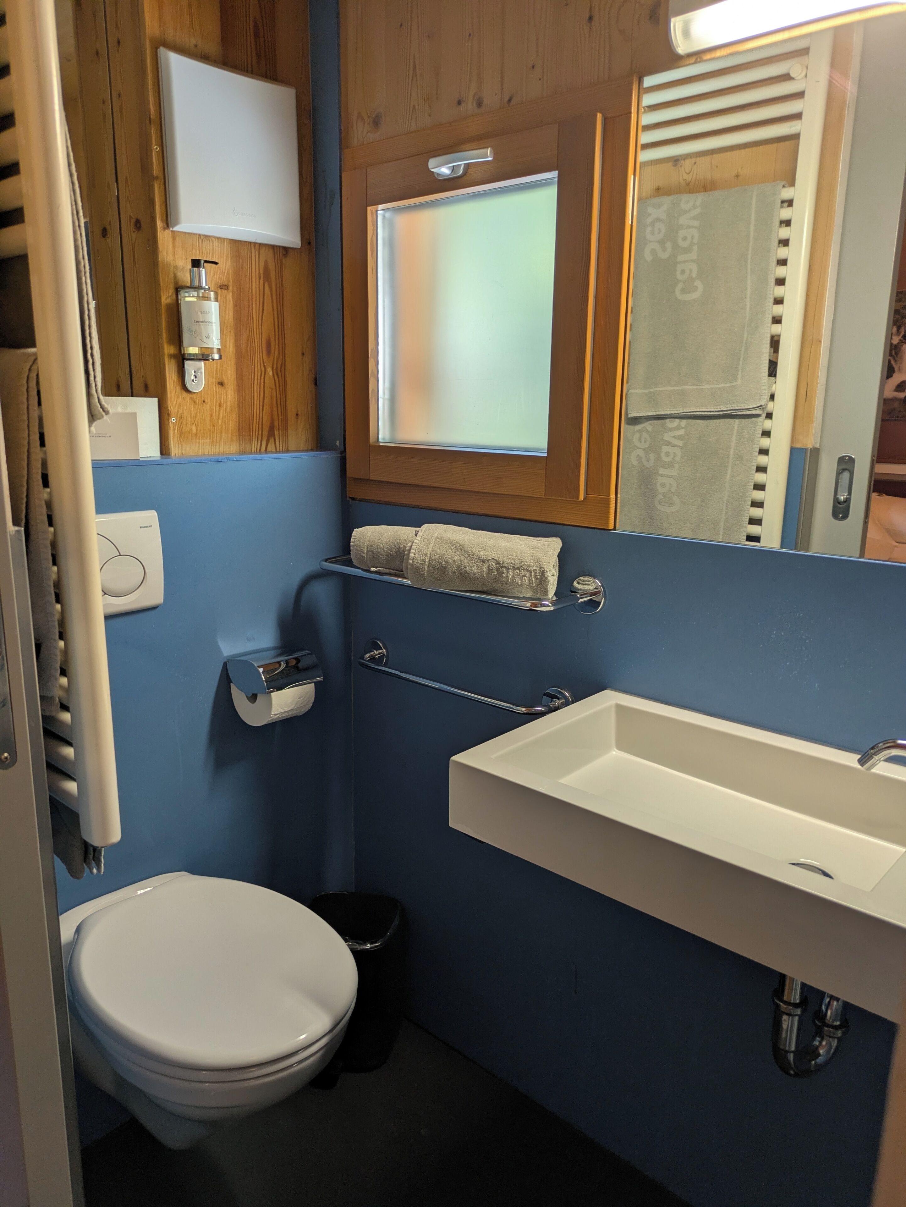 A compact bathroom with a white toilet, a sink, a towel rack, and a window with blinds.