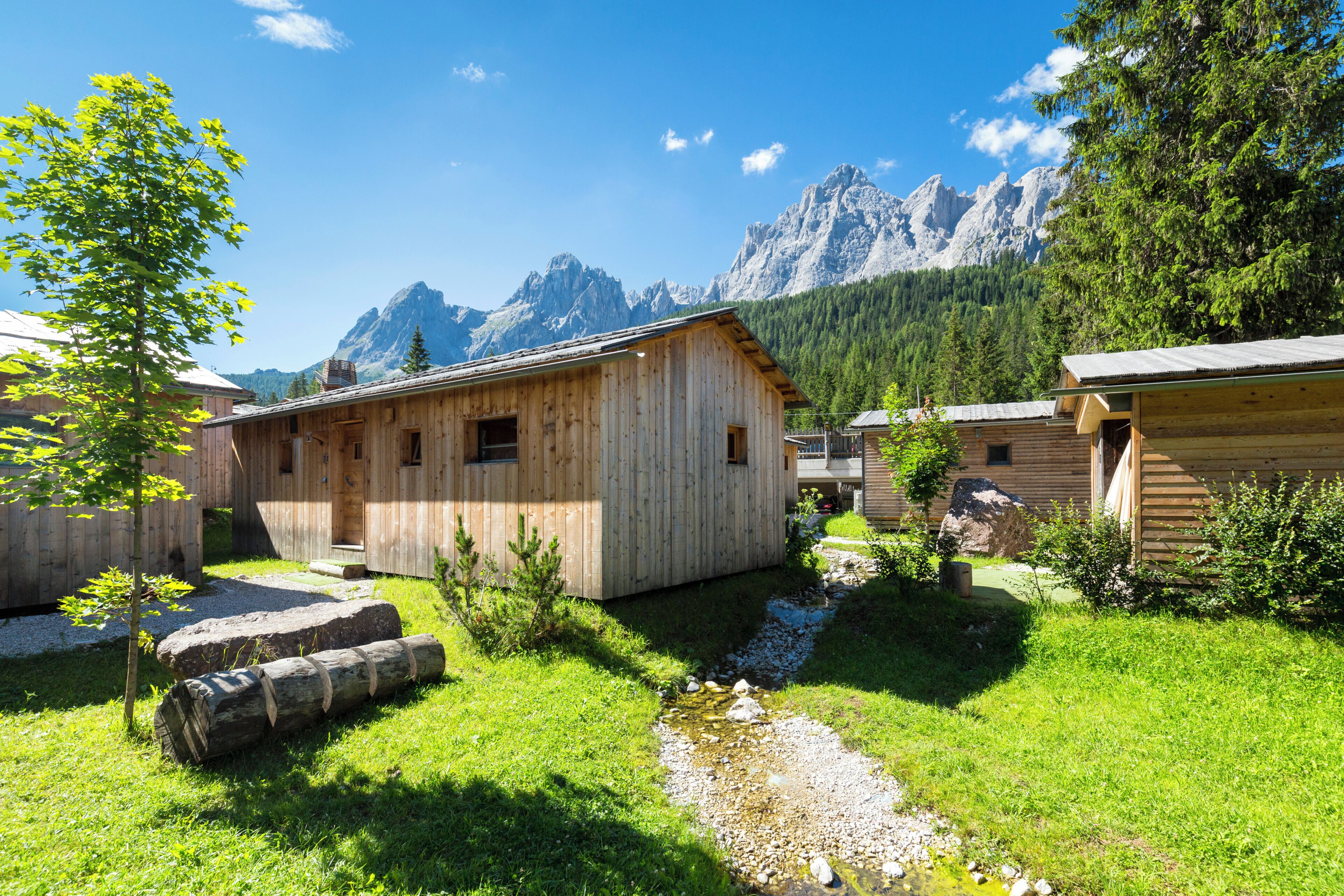 A wooden cabin with a small garden and a stream, surrounded by mountains.