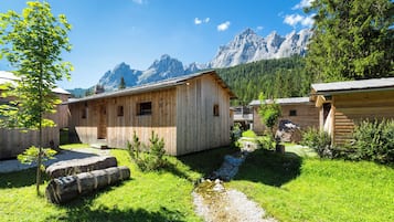 A wooden cabin with a small garden and a stream, surrounded by mountains.