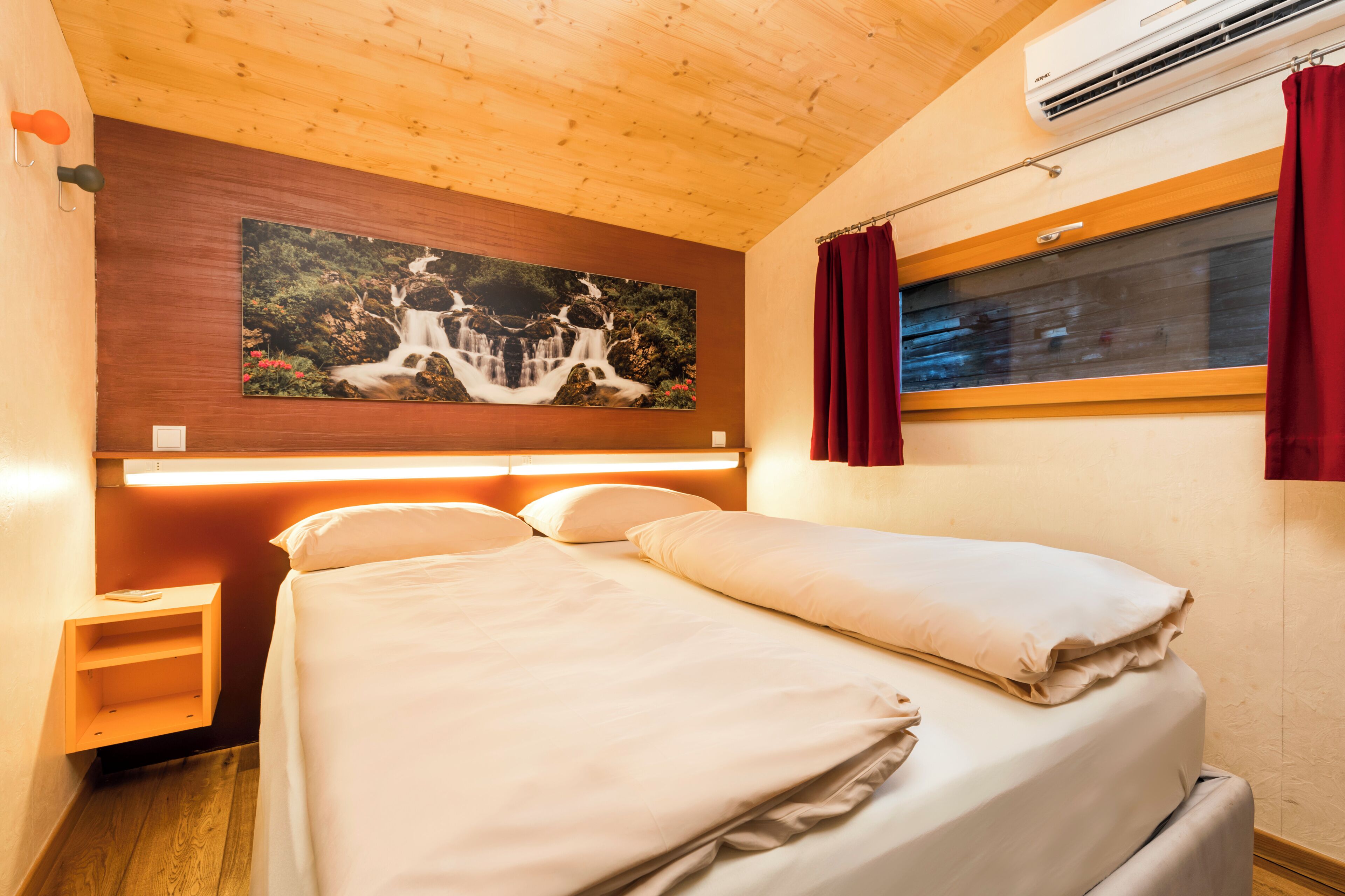 Une chambre à coucher avec un lit, une table de chevet, un climatiseur fixé au mur, une fenêtre avec des rideaux et un tableau au-dessus du lit.