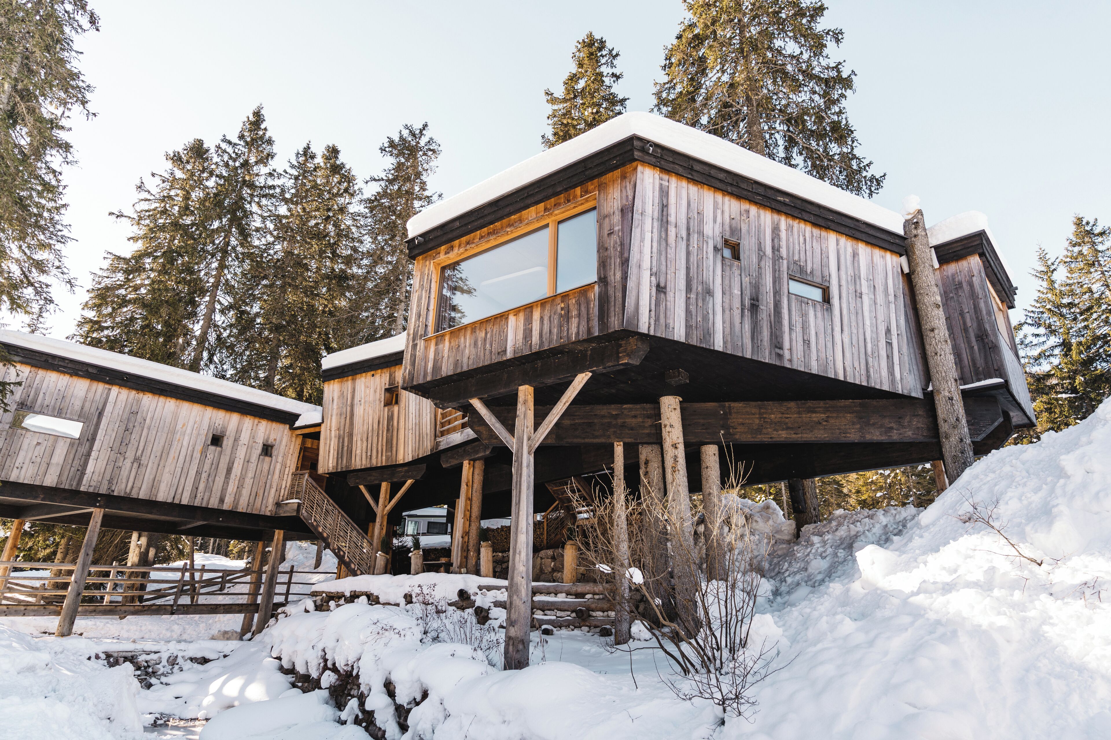 Eine Holzhütte mit großem Fenster, auf Stelzen stehend, umgeben von Schnee und Bäumen.