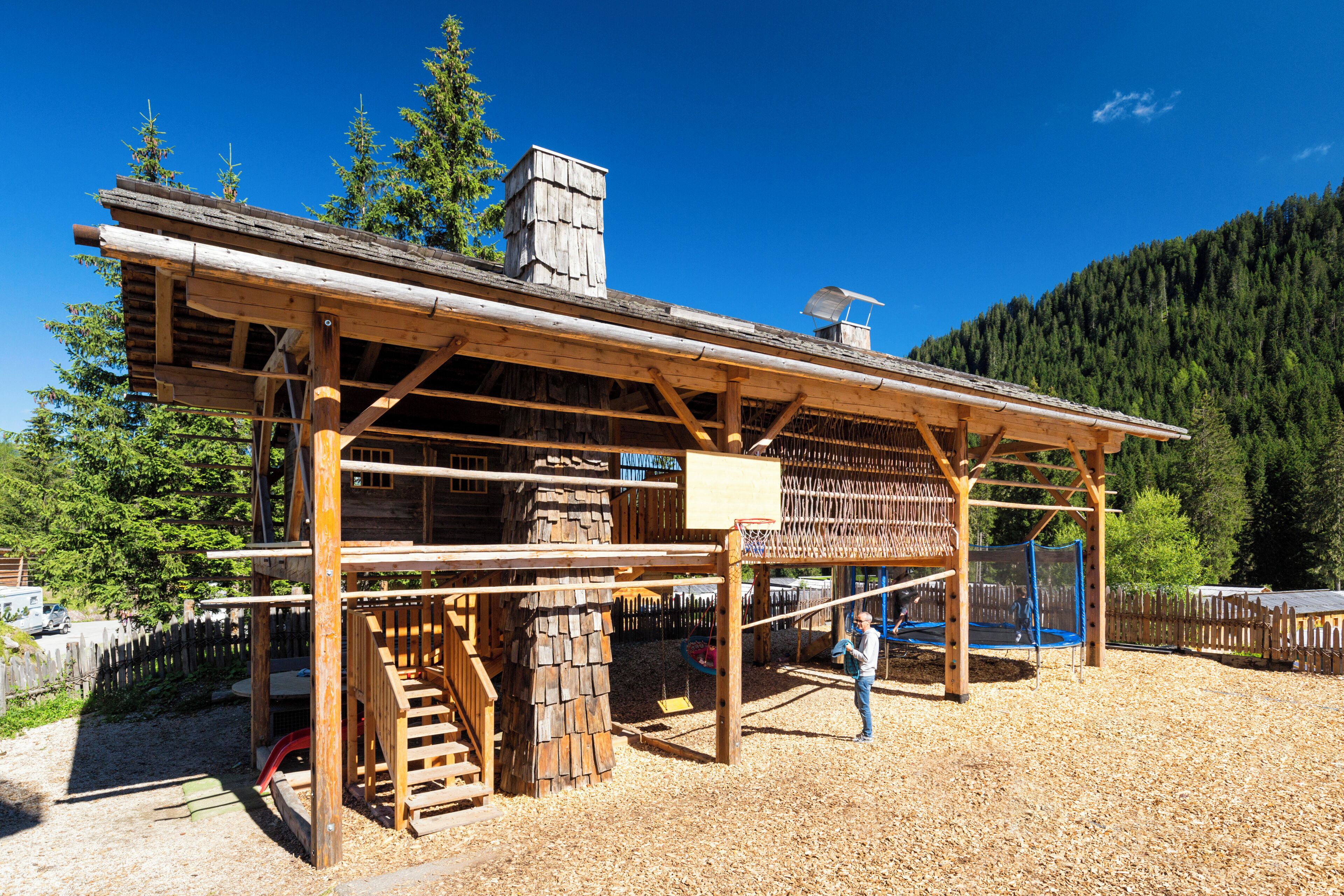 A rustic wooden structure with a covered porch, a chimney, and a playground area with a slide and climbing net.