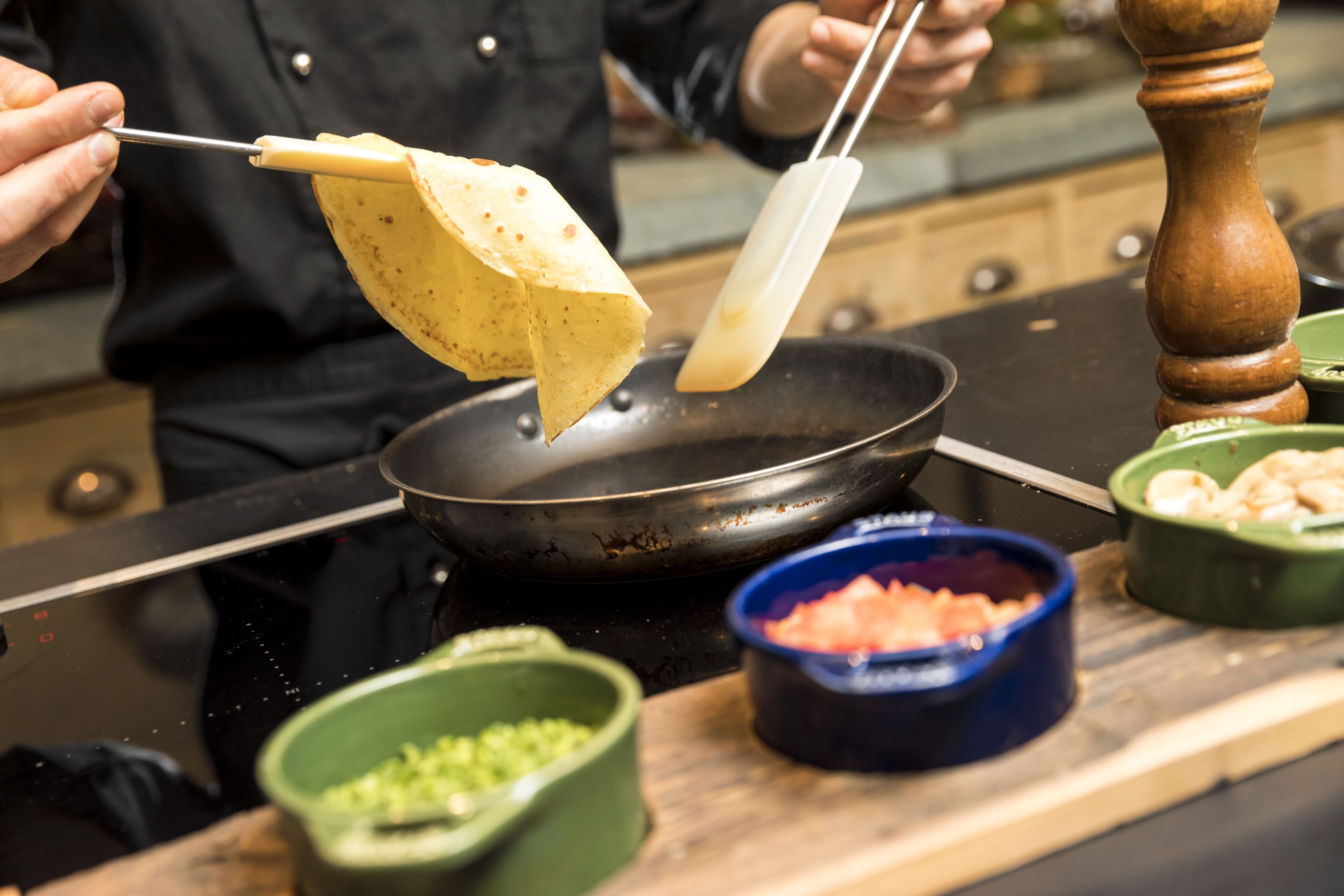 A person cooking with a frying pan on a stove, holding a tortilla with tongs.