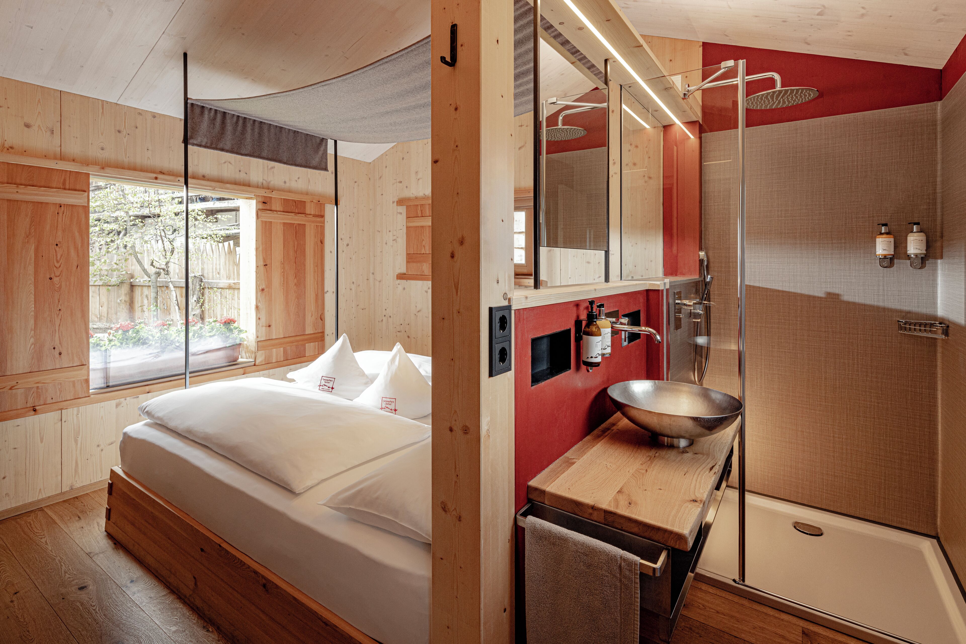 A modern bedroom with a large bed, a wooden floor, a window, and a bathroom with a shower, sink, and towel rack.