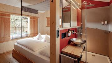 A modern bedroom with a large bed, a wooden floor, a window, and a bathroom with a shower, sink, and towel rack.
