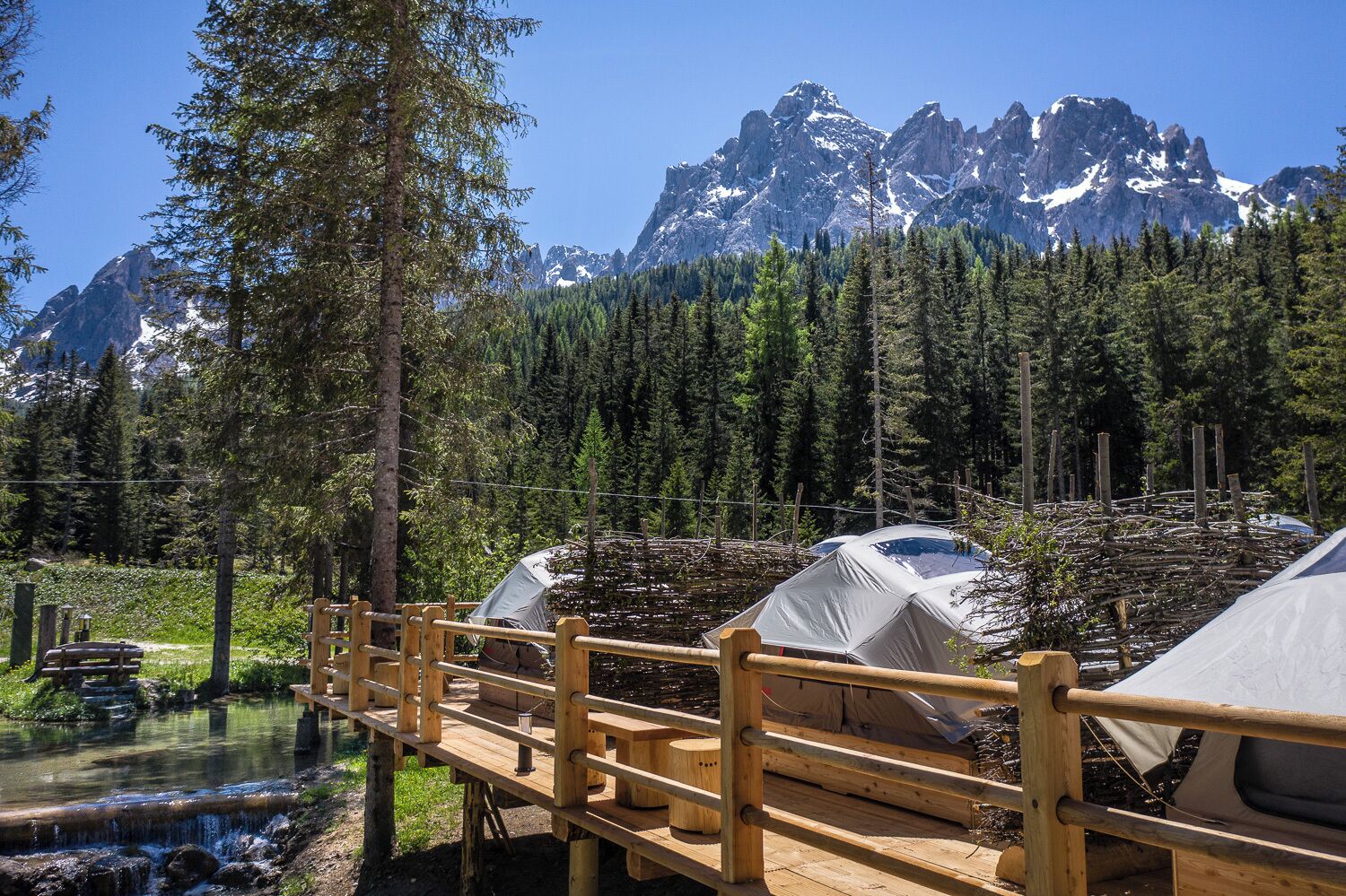 A wooden bridge leading to a campsite with tents, surrounded by a forest and a mountain range.