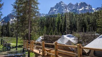 A wooden bridge leading to a campsite with tents, surrounded by a forest and a mountain range.