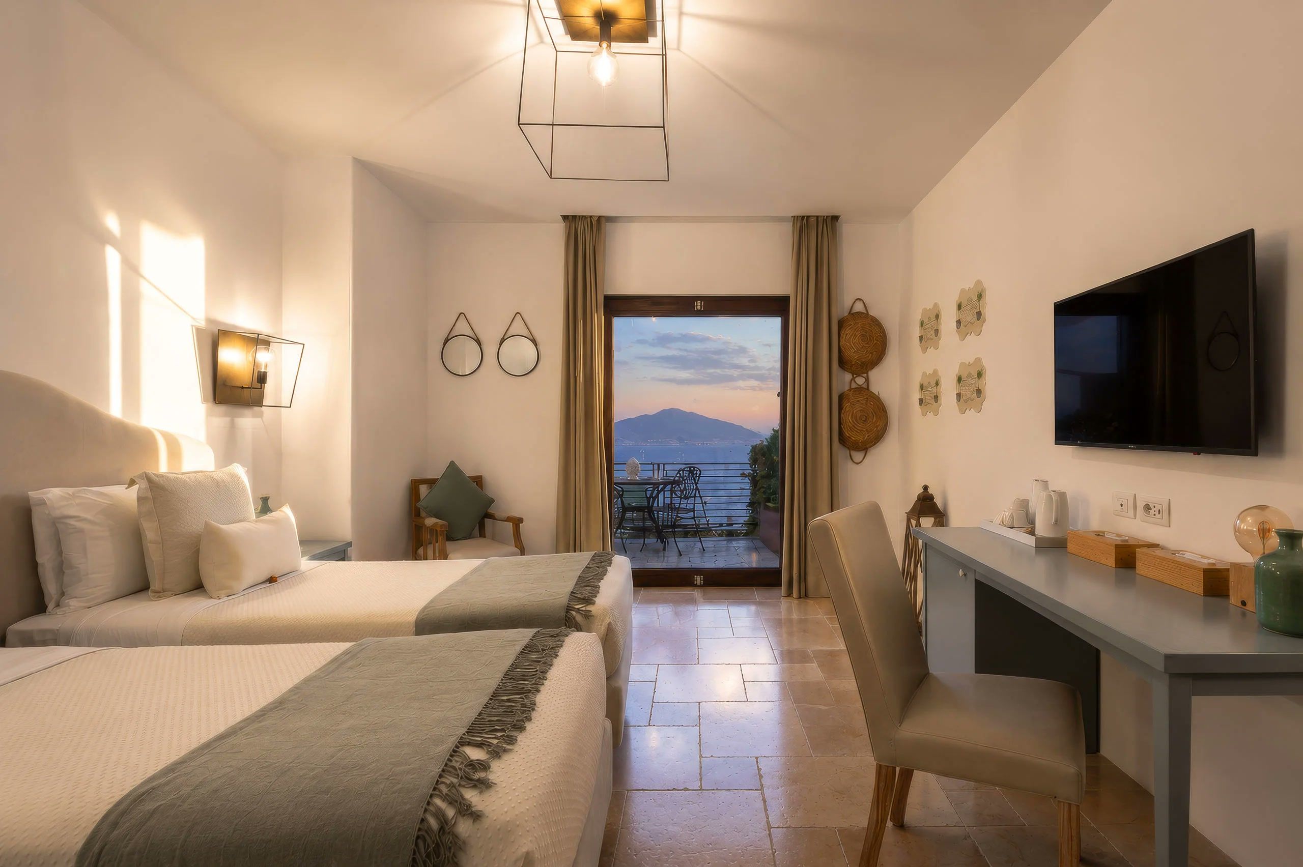 A hotel room with two beds, a desk, a television, and a balcony with a view.