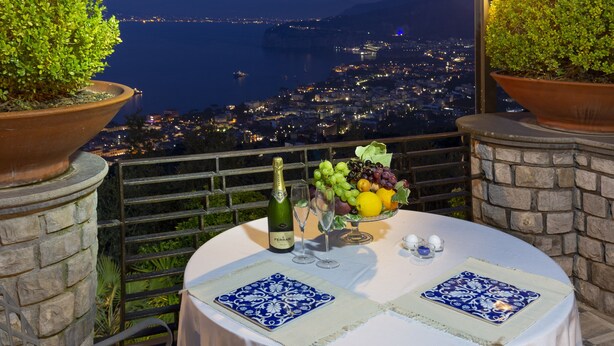 A table with a bottle of champagne, glasses, and a fruit bowl, overlooking a cityscape at night. - Romantic Relais Sorrento (Sorrento)