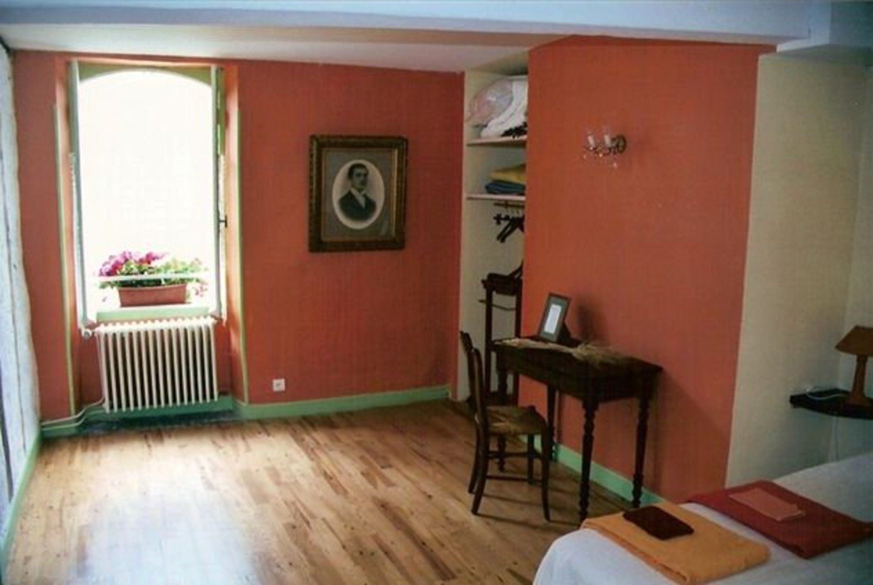 A room with a wooden floor, a bed with pillows, a chair, a desk with a chair, a radiator, and a window with a view of flowers.