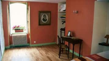 A room with a wooden floor, a bed with pillows, a chair, a desk with a chair, a radiator, and a window with a view of flowers.
