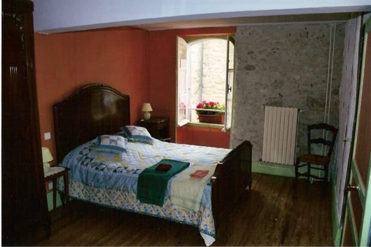 Een slaapkamer met een houten bed, een nachtkastje, een stoel, een raam met uitzicht op bloemen en een radiator.
