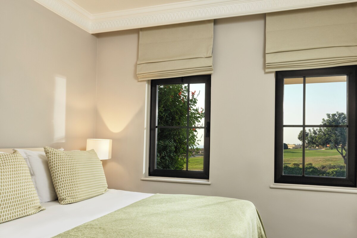 A bedroom with a bed, a nightstand, a lamp, and two windows with views of greenery.