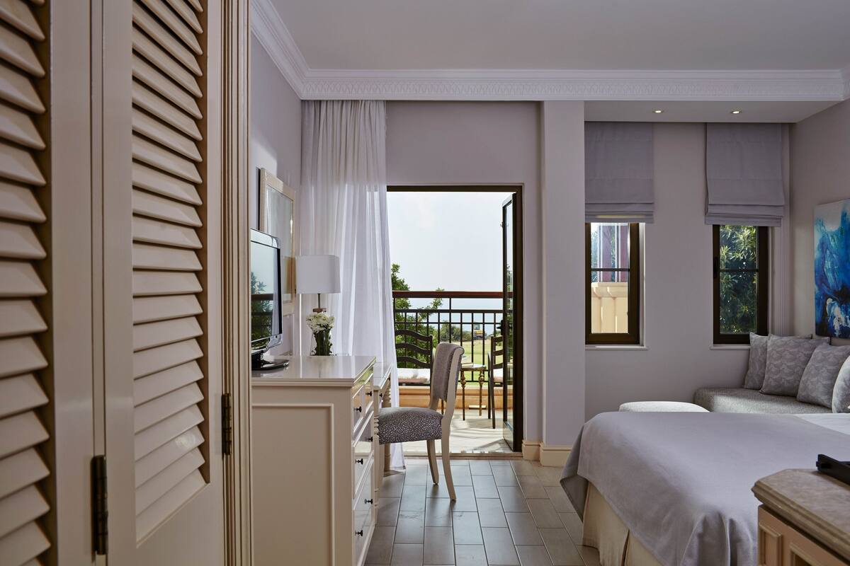 A bedroom with a bed, a desk with a chair, a balcony with a view, and a window with a painting.