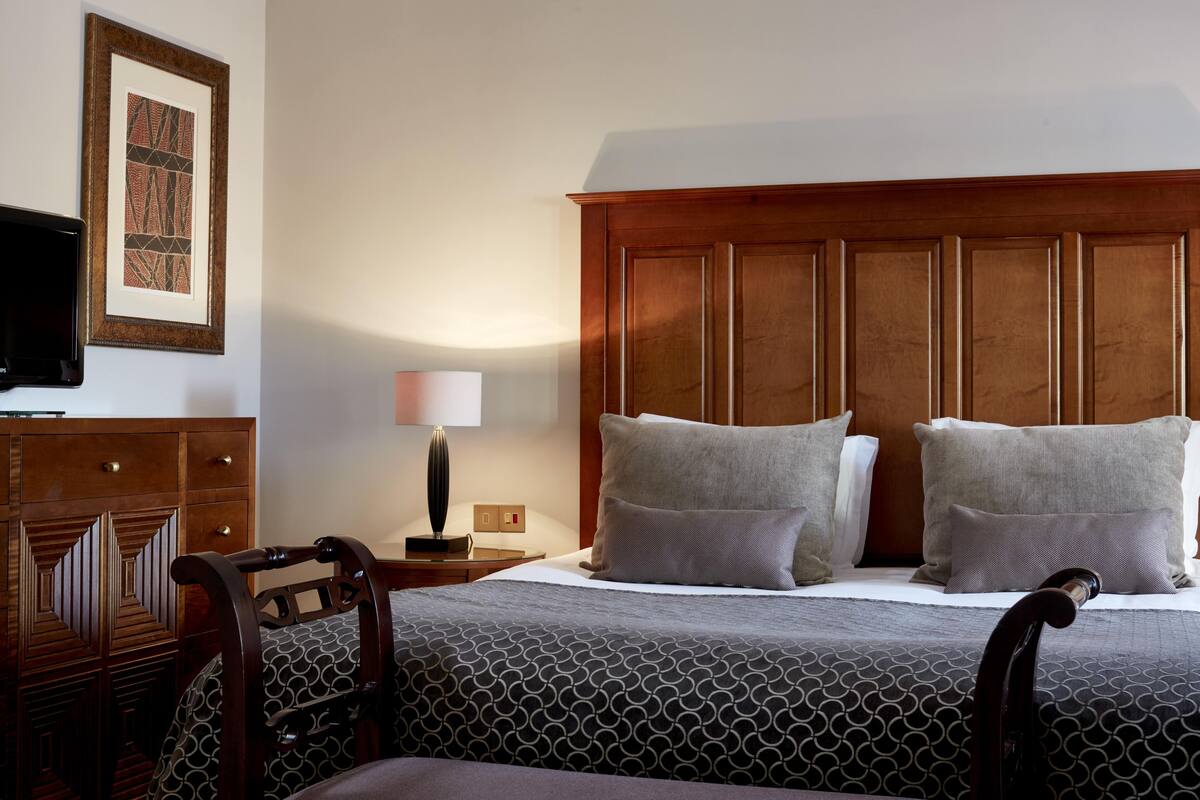 A hotel room with a wooden headboard, two pillows, a bedside table with a lamp, and a TV mounted on the wall.