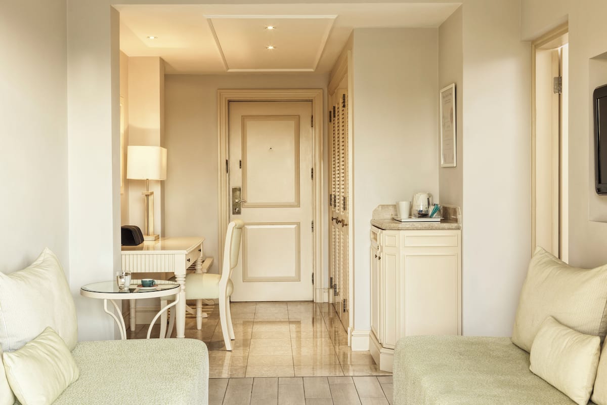A hotel room with a seating area, a small table with a lamp, a coffee maker, and a cabinet.