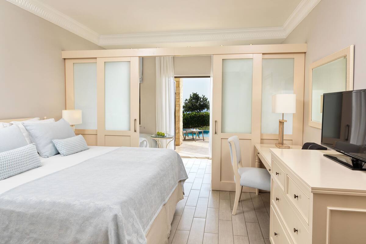 A hotel room with a bed, a desk, a chair, a TV, and a view of the outdoor pool area.