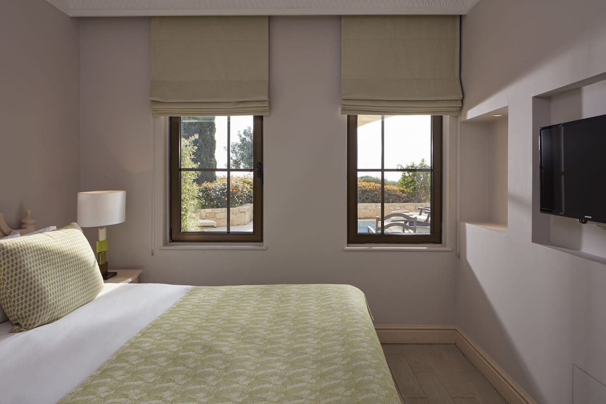 A hotel room with a bed, two windows with blinds, a TV mounted on the wall, and a view of an outdoor area.