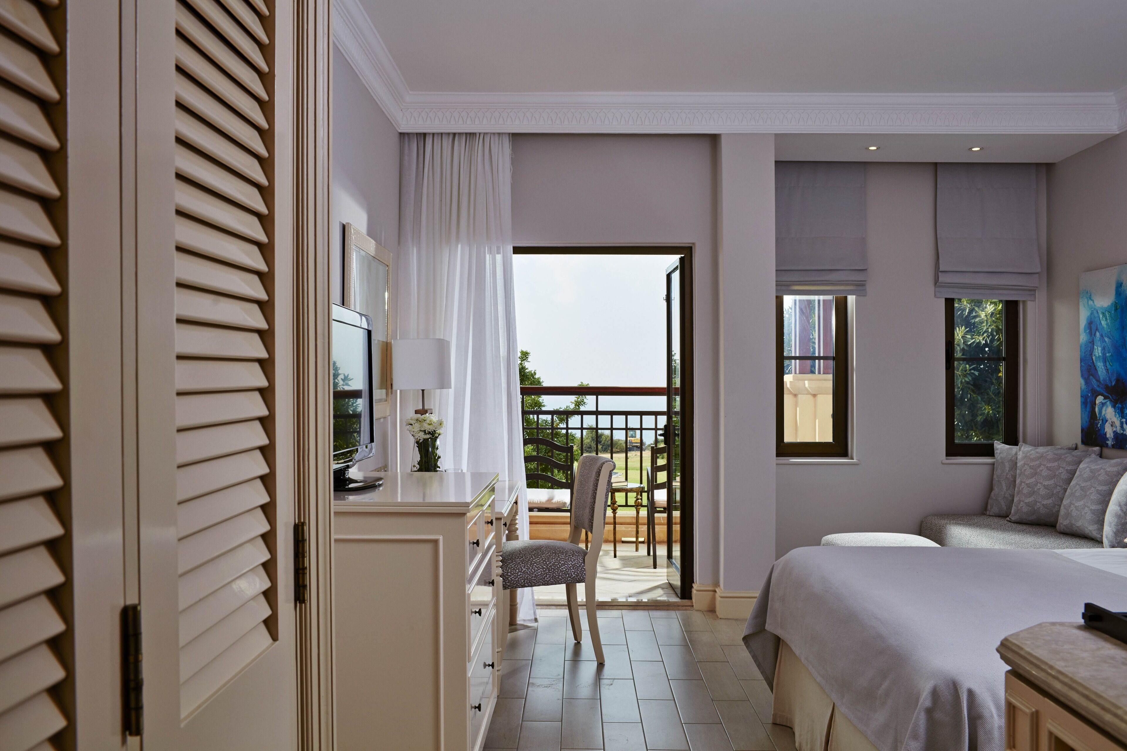 Ein Schlafzimmer mit Bett, Schreibtisch mit Stuhl, einem Balkon mit Aussicht und einem Fenster mit einem Gemälde.
