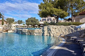 Área de piscina com espreguiçadeiras, borda rochosa e um edifício ao fundo.