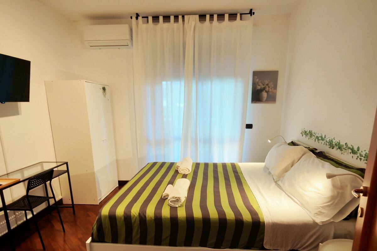 A bedroom with a bed, a desk, a chair, a television, and a window with curtains.