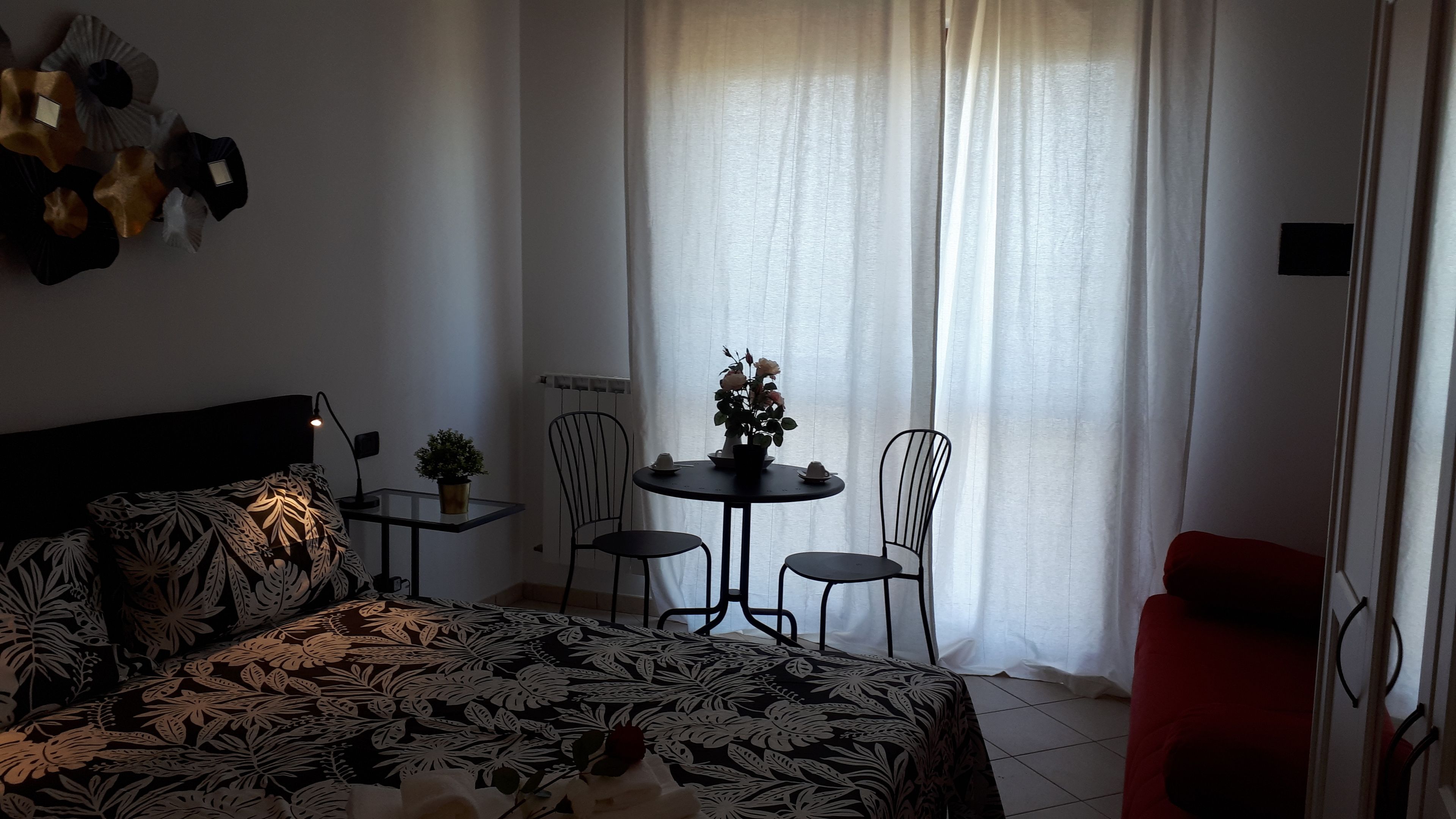 Quarto com cama, uma mesinha com cadeiras, uma planta em vaso e janela com cortinas.