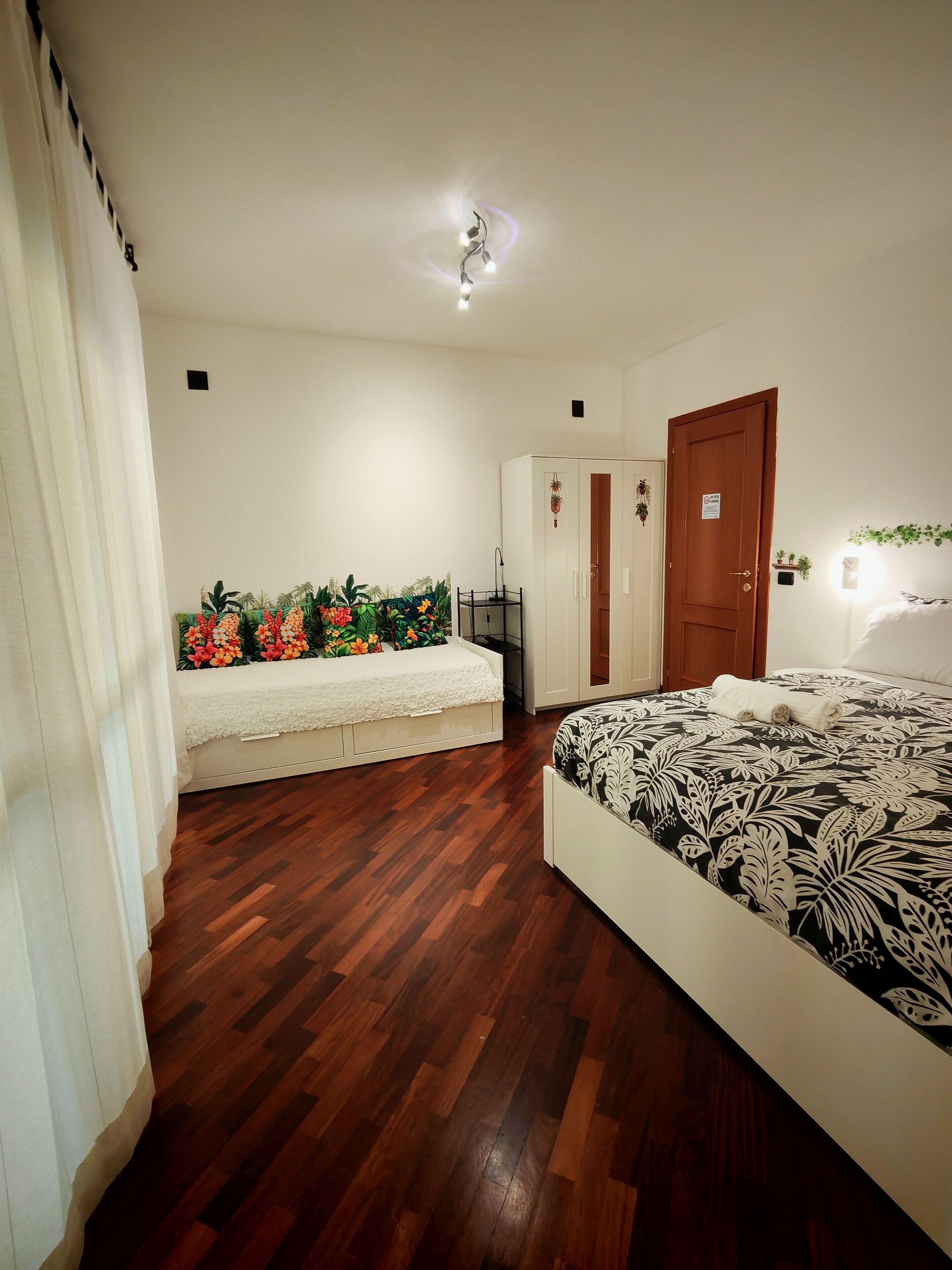 A bedroom with a wooden floor, a bed with a patterned bedspread, a white bedside table, a wardrobe, and a decorative wall hanging.