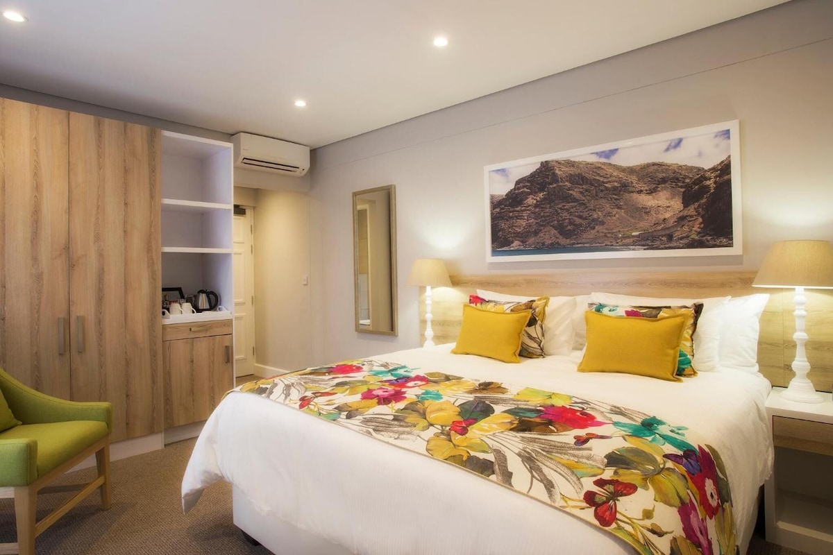 A hotel room with a bed, a green chair, a wardrobe, a mirror, a painting, and a nightstand with a lamp.