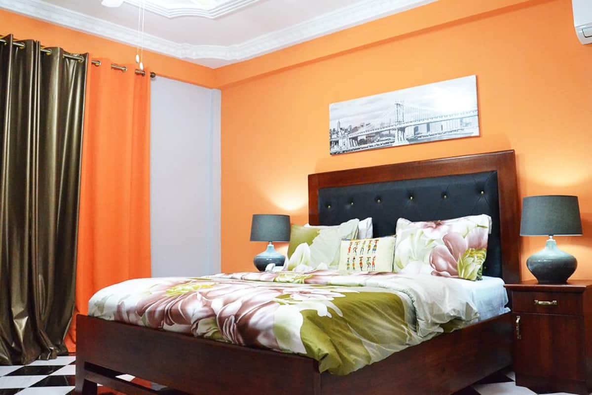 A bedroom with an orange wall, a bed with floral bedding, two bedside lamps, and a picture of a bridge.
