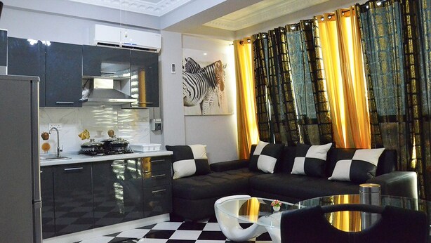 A modern kitchen with black cabinets, a black and white checkered floor, and a black sofa with white cushions. - Royal Haveala Apartments (Accra)