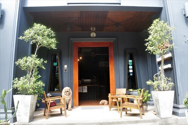 A building entrance with a wooden door, two potted plants, a lion statue, and a small table and chair set outside.
