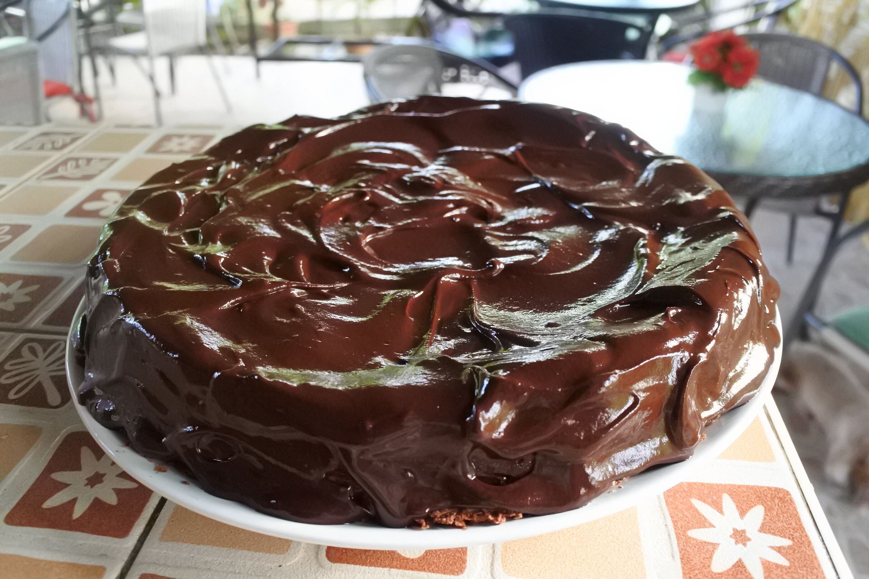 A chocolate cake on a white plate on a table with a patterned tablecloth.