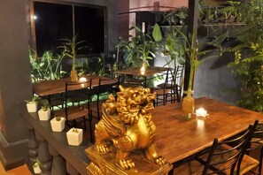 A dining area with a wooden table, chairs, a golden lion statue, potted plants, and a window overlooking a garden. - Hi Karon Beach Hotel (Karon)
