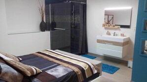 Een moderne slaapkamer met een glazen douche, een nachtkastje, een bed met gestreepte lakens, een witte ladekast en een spiegel boven de ladekast.