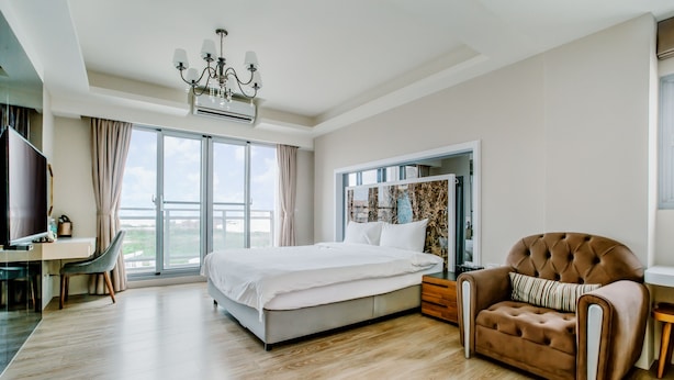Una habitación de hotel moderna con una cama grande, un área de descanso con un sillón marrón, un escritorio y un ventanal con vistas.