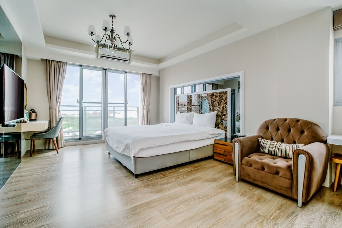 A modern hotel room with a large bed, a sitting area with a brown armchair, a desk, and a large window offering a view.
