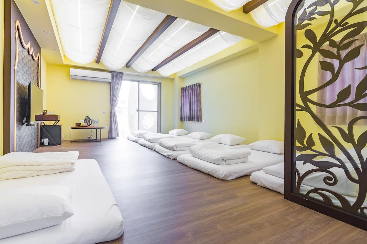 A room with a wooden floor, a bed with white bedding, a massage table with white cushions, a small table with a plant, and a decorative partition with leaf patterns.