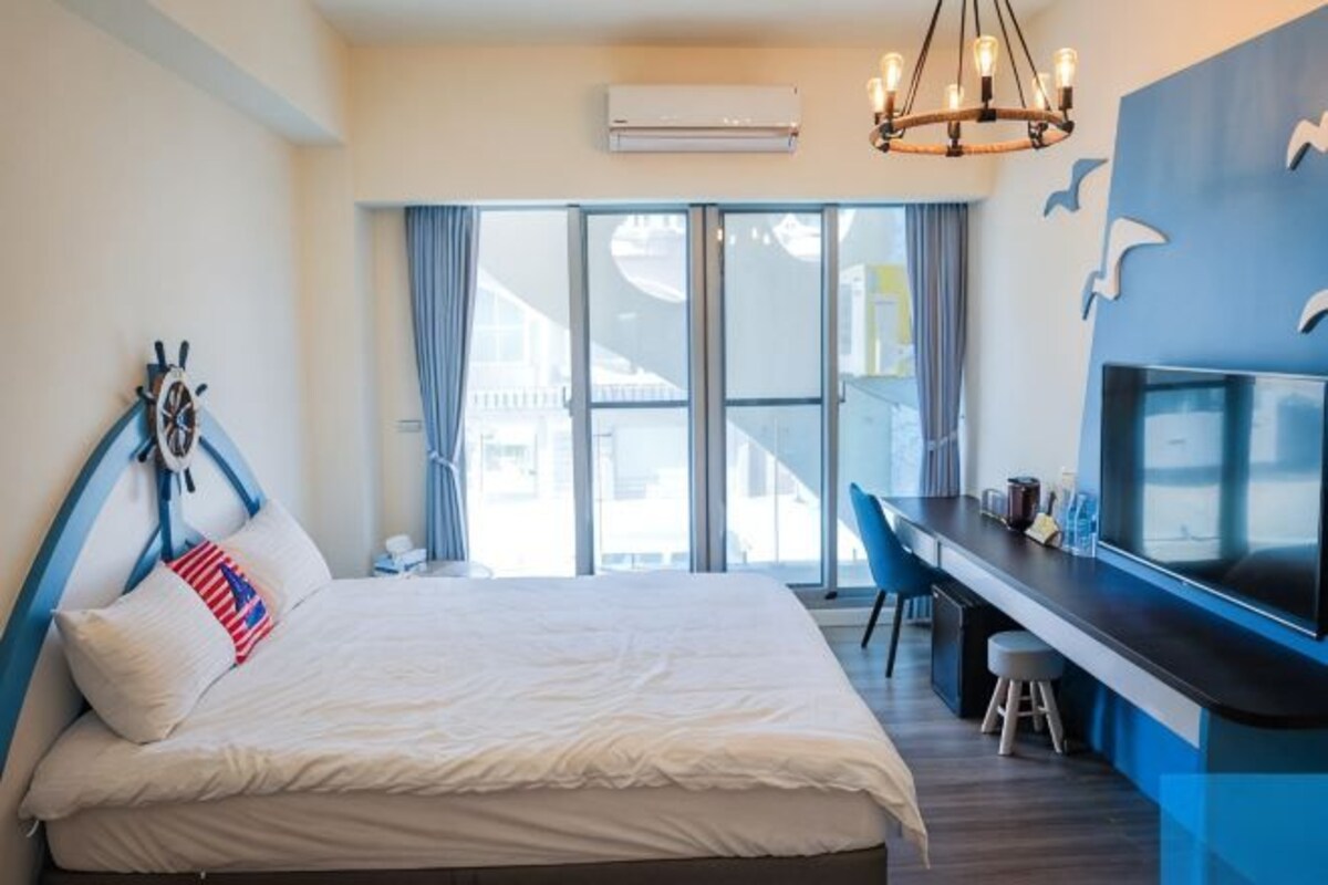 A hotel room with a bed, a desk, a chair, an air conditioning unit, and a view through a sliding door.