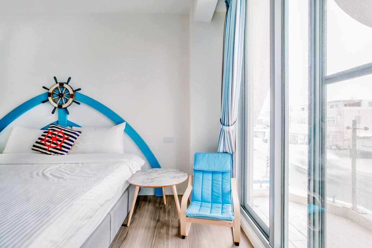 A modern hotel room with a bed, a small round table, a blue chair, and a large window offering a city view.