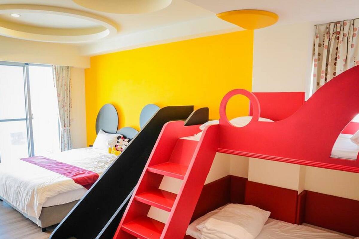 A hotel room with a bed, a red and white bunk bed, and a large window.