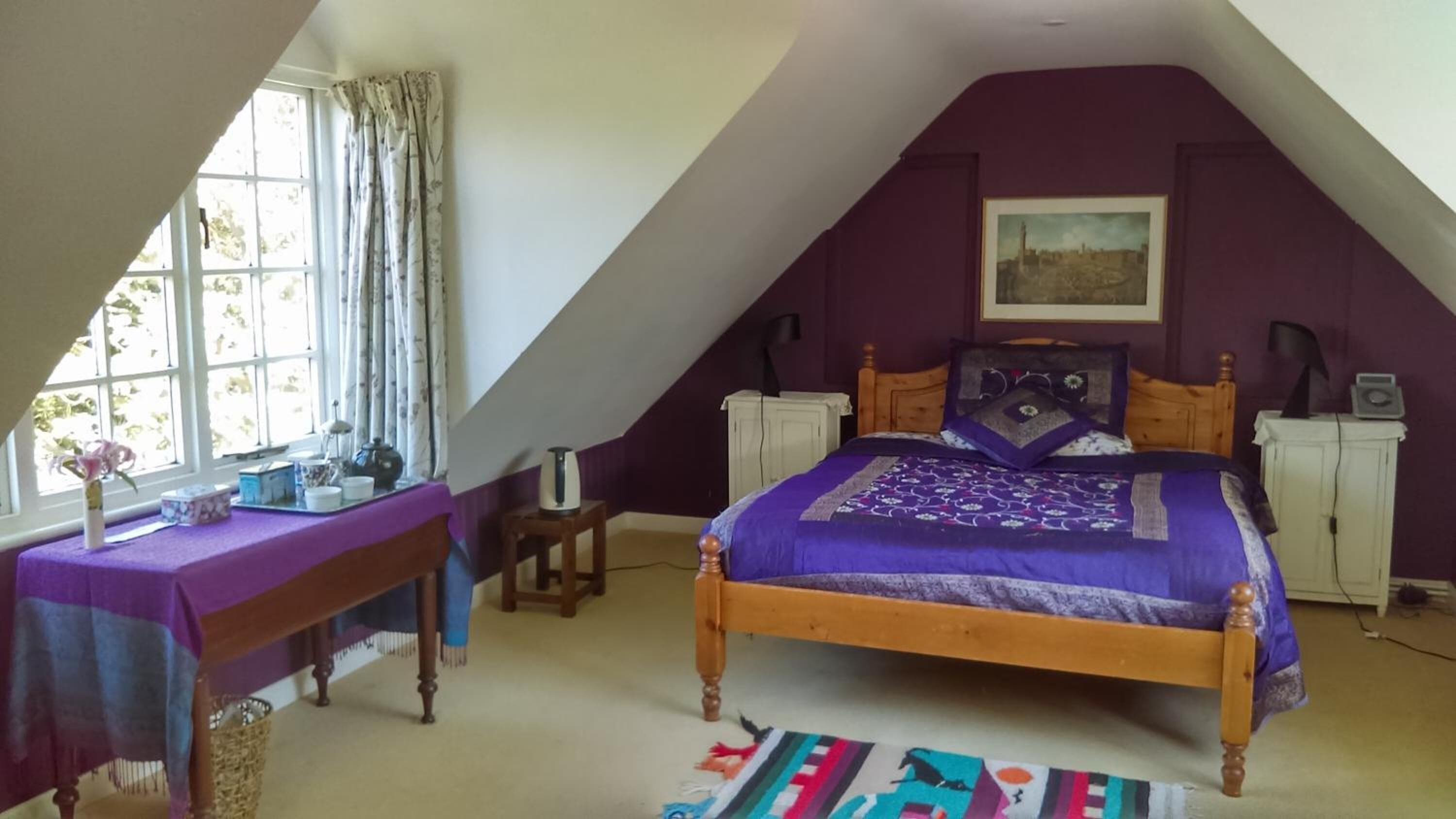 Un dormitorio con techo inclinado, una cama con colcha morada, una mesita con mantel morado, una silla y un ventanal con cortinas.