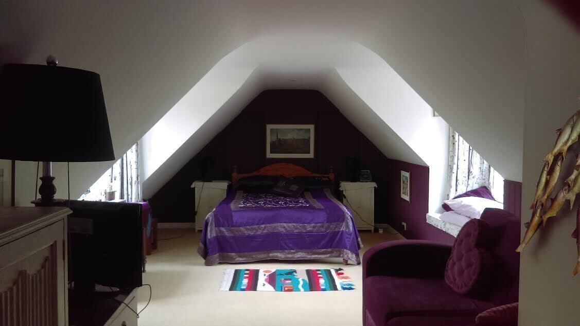 Un dormitorio con techo inclinado, una cama con colcha morada, un sillón morado y una alfombra colorida.
