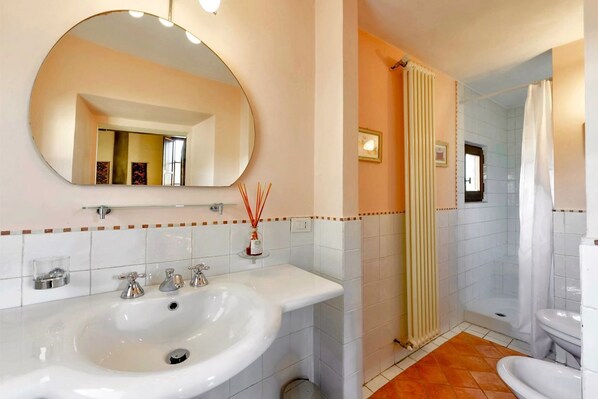 A bathroom with a large oval mirror, a sink with three faucets, a shower area, and a toilet.